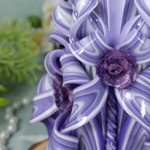 Puede incluir: Una vela tallada a mano de color morado y blanco con un dise&ntilde;o de espiral y tres flores moradas. La vela est&aacute; sobre una base de madera.