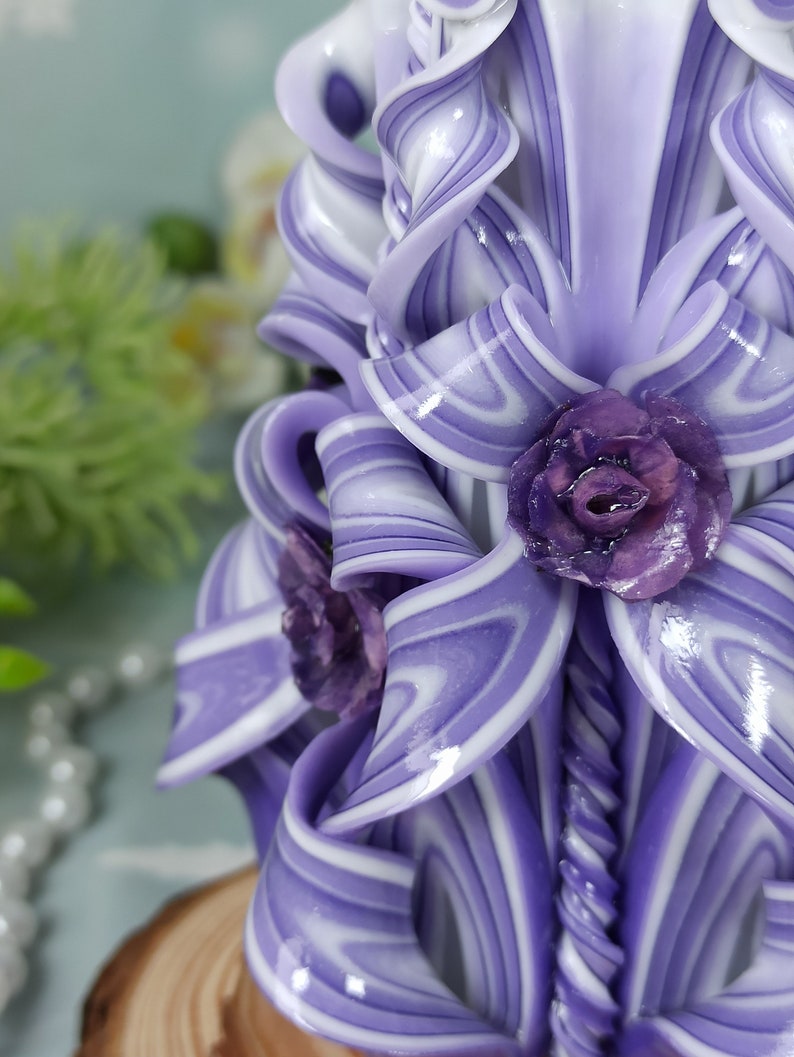 Puede incluir: Una vela tallada a mano de color morado y blanco con un dise&ntilde;o de espiral y tres flores moradas. La vela est&aacute; sobre una base de madera.