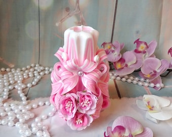 May include: A pink and white striped candle with a floral design. The candle is decorated with pink flowers and a white bow. The candle is sitting on a white surface with a pearl necklace and pink flowers.