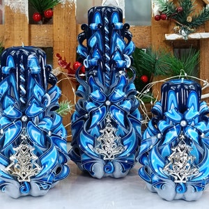 Puede incluir: Tres velas talladas azules y blancas con adornos de árbol de Navidad plateados. Las velas están decoradas con cintas azules y blancas y perlas.