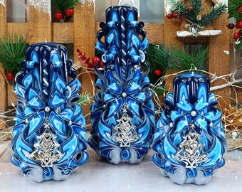 May include: Three blue and white carved candles with silver Christmas tree ornaments. The candles are decorated with blue and white ribbons and pearls.
