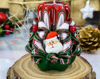 May include: A red and green Christmas candle with a Santa Claus face in the center. The candle is decorated with white and red stripes and green leaves.