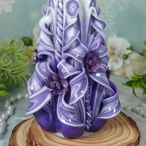 Puede incluir: Una vela tallada a mano de color morado y blanco con un dise&ntilde;o de espiral y detalles florales. La vela est&aacute; sobre una base de madera.