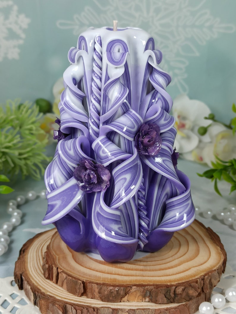 Puede incluir: Una vela tallada a mano de color morado y blanco con un dise&ntilde;o de espiral y detalles florales. La vela est&aacute; sobre una base de madera.