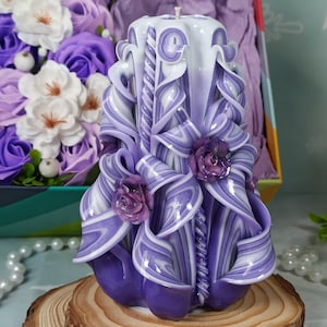 Puede incluir: Una vela tallada de color morado y blanco con un dise&ntilde;o de espiral y tres rosas moradas. La vela est&aacute; sentada sobre una base de madera.