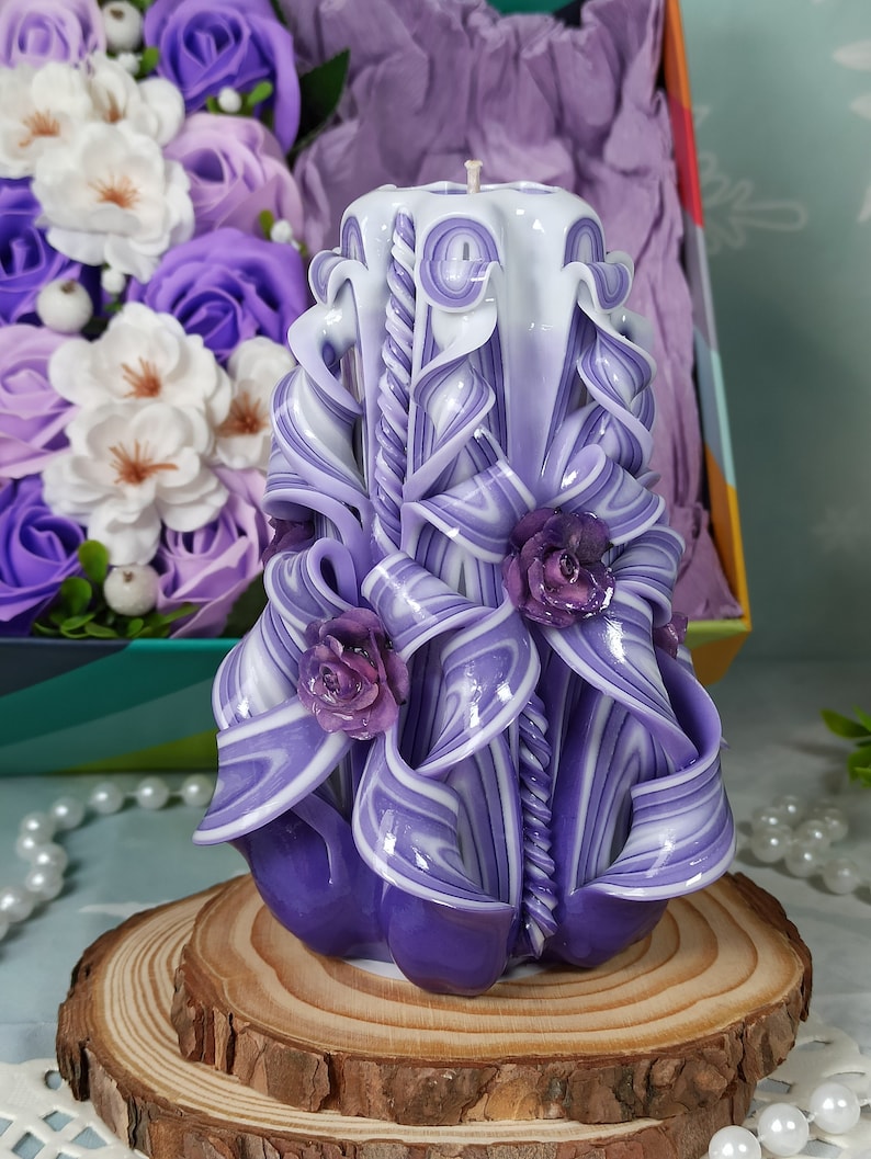 Puede incluir: Una vela tallada de color morado y blanco con un dise&ntilde;o de espiral y tres rosas moradas. La vela est&aacute; sentada sobre una base de madera.