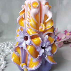 May include: A hand-carved candle with a purple, yellow, and pink striped design. The candle is decorated with three purple flowers.