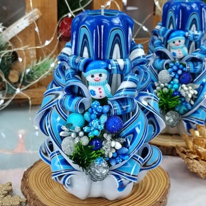 以下が含まれることがあります： 雪だるまの置物と青と銀の装飾が施された青と白のストライプのキャンドル2本。キャンドルは木製の台座に乗っています。