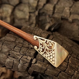 Puede incluir: Un palillo de pelo de madera con un extremo decorativo de metal dorado. El extremo metálico presenta un diseño calado ornamentado y una base rectangular nacarada. El accesorio para el cabello se encuentra sobre un fondo oscuro y texturizado.