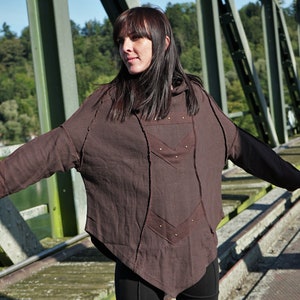 May include: A brown hooded tunic with a unique design featuring multiple seams and stitching. The tunic is worn by a person standing on a bridge.