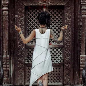 May include: A woman with tattoos stands in front of a carved wooden door with two stone lion statues on either side. She is wearing a white sleeveless dress and has her arms outstretched.