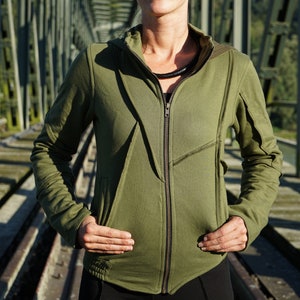 May include: A woman wearing a green hooded zip-up jacket with a black necklace. The jacket has a unique design with angled seams and a zip-up front.