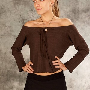 Può includere: Una donna indossa una blusa marrone scollata con una minigonna nera e stivali neri. È in piedi davanti a uno sfondo grigio.