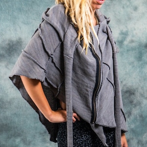 Könnte beinhalten: Eine Frau mit langen blonden Dreadlocks trägt einen grauen Kapuzenpullover mit Reißverschluss und ausgefranstem Saum. Der Pullover hat an jeder Seite einen langen Quasten. Sie trägt eine schwarze Hose mit einem weißen Muster.