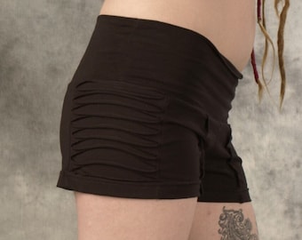 May include: A pair of brown shorts with a gathered detail on the front. The shorts are worn by a person with tattoos on their legs and are paired with brown boots.