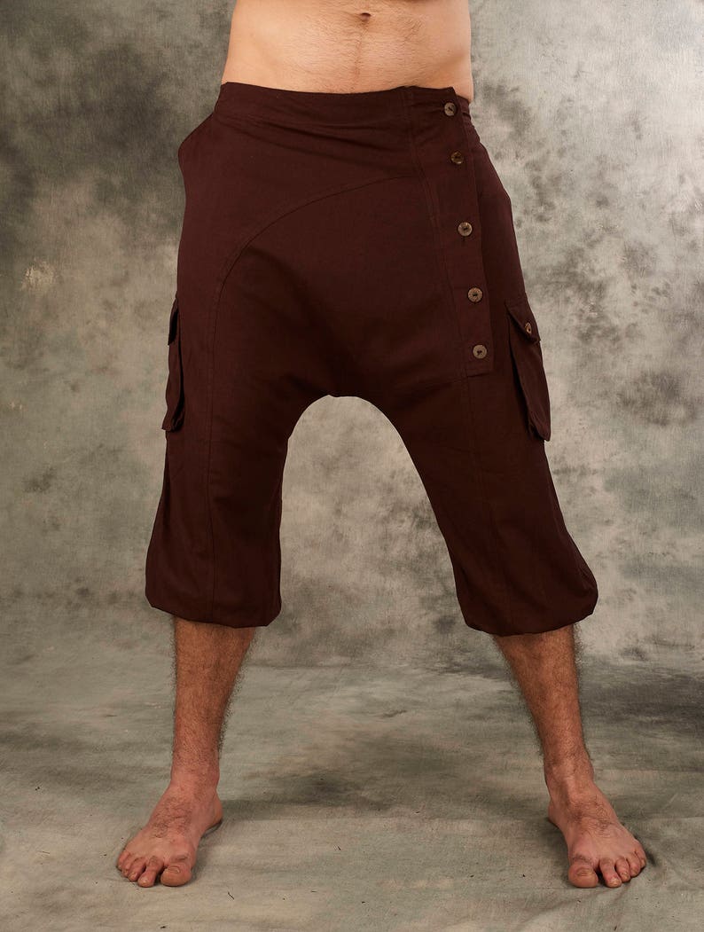 May include: Dark brown drop-crotch pants featuring a high-waisted design with a side button closure. These cropped trousers include two side cargo pockets with flaps and a smooth, solid-colored fabric construction that ends below the knees.