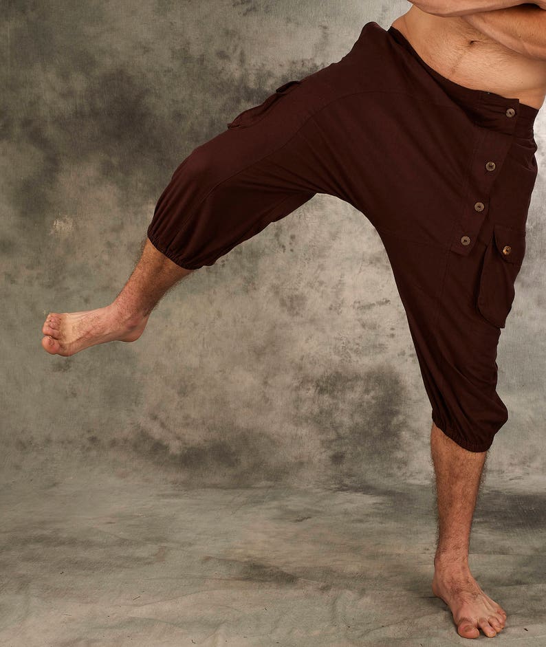 May include: Dark brown cropped pants featuring a drop-crotch design, an asymmetrical button-down waistband closure, and side cargo pockets. The leg openings are gathered with elastic, creating a tapered fit that ends below the knee.