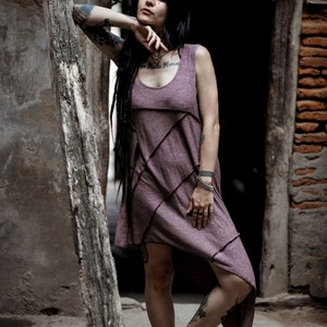 Peut inclure: Une femme aux longs cheveux noirs et tatouée porte une robe asymétrique violette avec un design unique. Elle se tient devant un mur de briques avec une porte en bois.