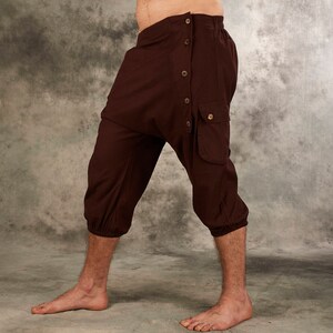 May include: Dark brown cropped pants featuring a drop-crotch design, an elasticized waistband, and a side panel secured with five buttons. The pants include a single flap pocket on the side and gathered elastic cuffs at the bottom of each leg.