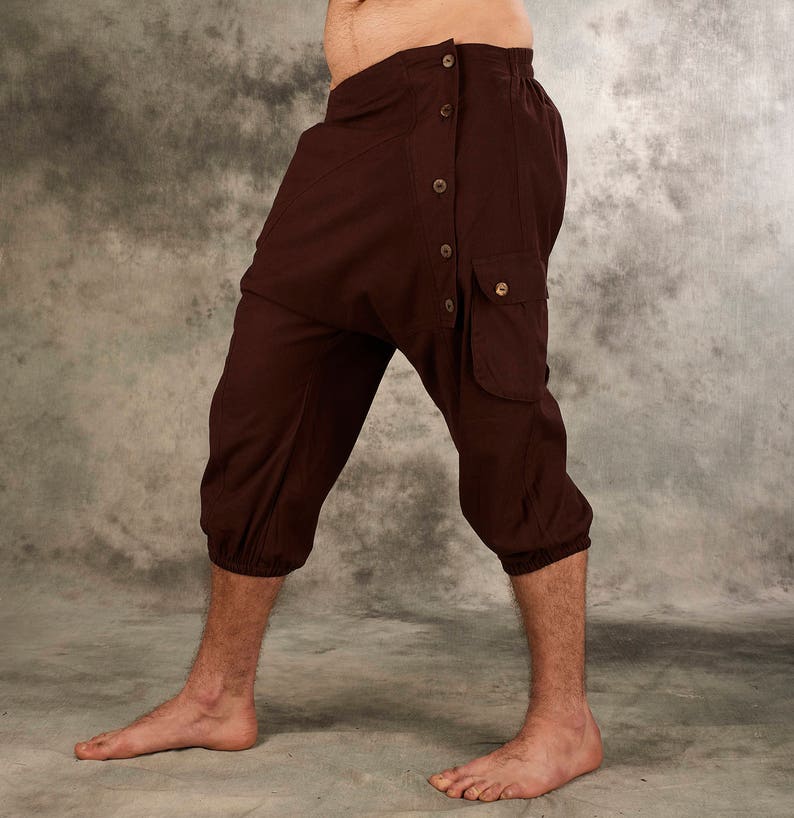 May include: Dark brown cropped pants featuring a drop-crotch design, an elasticized waistband, and a side panel secured with five buttons. The pants include a single flap pocket on the side and gathered elastic cuffs at the bottom of each leg.