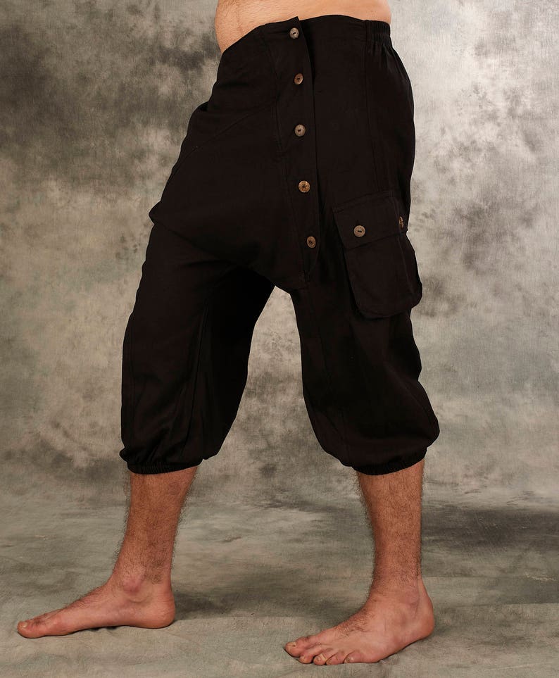 May include: Black cropped trousers featuring a drop-crotch design, an elasticized waistband, and a side panel secured with five light brown buttons. The garment includes a flap pocket on the side and elasticized cuffs at the bottom of each leg.