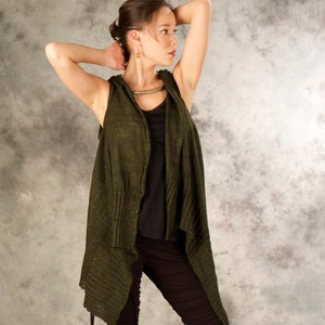 May include: A woman wearing a green knitted cardigan with a draped front and a black tank top underneath. She is standing against a grey background.