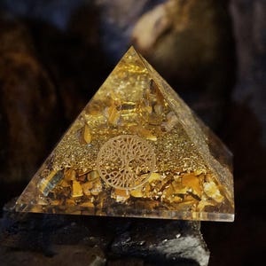 May include: A gold pyramid with a tree of life design inside. The pyramid is made of clear resin and filled with small pieces of gold and brown stones.