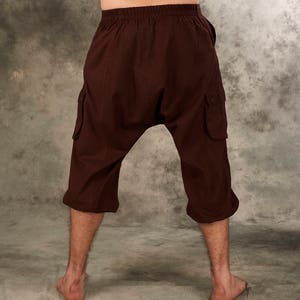 May include: Dark brown drop-crotch cargo pants featuring an elasticized waistband, two side cargo pockets with button closures, and a cropped length that ends below the knees. The pants have a loose, relaxed fit with a low-hanging crotch area.