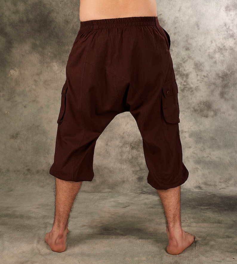 May include: Dark brown drop-crotch cargo pants featuring an elasticized waistband, two side cargo pockets with button closures, and a cropped length that ends below the knees. The pants have a loose, relaxed fit with a low-hanging crotch area.