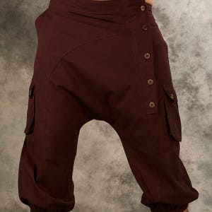 May include: Dark brown cropped pants featuring a drop-crotch design, an asymmetrical button-down front closure with five visible buttons, and two side cargo pockets with flaps. The leg openings are finished with elasticized cuffs.