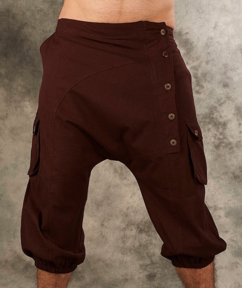 May include: Dark brown cropped pants featuring a drop-crotch design, an asymmetrical button-down front closure with five visible buttons, and two side cargo pockets with flaps. The leg openings are finished with elasticized cuffs.