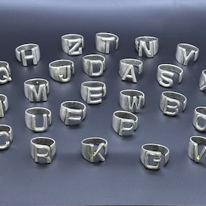 May include: A set of 26 silver rings, each featuring a different letter of the alphabet. The rings are all the same size and style, and they are arranged in a circle on a gray surface.