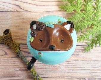 May include: A blue bicycle bell with a brown fox face sculpted on the front. The fox has a white nose and black eyes. The fox is wearing a wreath of green leaves and brown pinecones.