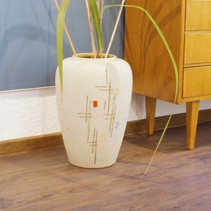 Könnte beinhalten: Eine weiße Keramikvase mit geometrischem Muster in Gelb-, Rot- und Blautönen. Die Vase ist mit getrockneten Schilf gefüllt und steht auf einem Holzboden.