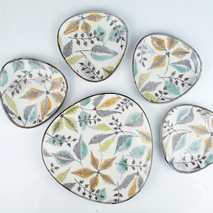 Pode incluir: Um conjunto de seis pratos de cerâmica com um padrão de folhas. Os pratos são de vários tamanhos e formas, com fundo branco e um desenho de folhas cinzentas, amarelas e verde-azuladas. As bordas são castanho-escuras.