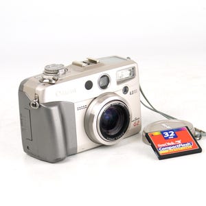 Può includere: Una fotocamera digitale Canon PowerShot G2 color argento e grigio con obiettivo da 4,0 megapixel, copriobiettivo e scheda di memoria SanDisk CompactFlash da 32 MB. La fotocamera presenta un obiettivo zoom, flash integrato e ghiere.