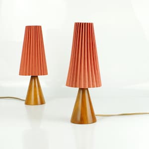 May include: Two matching table lamps. Each lamp has a cone-shaped wooden base and a pleated, coral-colored shade. The lamps are connected to a power source with brown cords. The lamps are on a white surface.