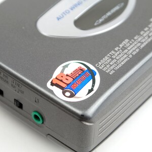 May include: A gray portable cassette player with a silver top panel labeled AUTO WIND and AUTO REVERSE. A circular sticker displays 18 HOURS LONG BATTERY LIFE with a blue battery graphic. The side features a green headphone jack and black switches.