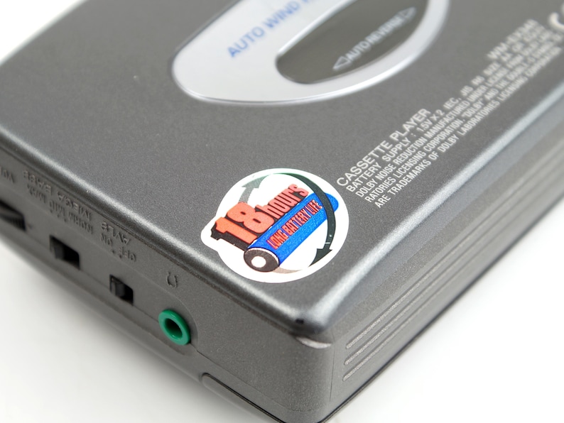 May include: A gray portable cassette player with a silver top panel labeled AUTO WIND and AUTO REVERSE. A circular sticker displays 18 HOURS LONG BATTERY LIFE with a blue battery graphic. The side features a green headphone jack and black switches.