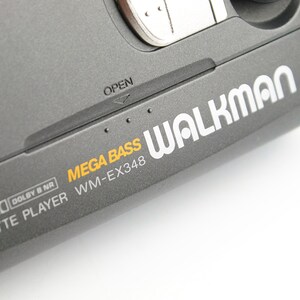 May include: A dark gray portable cassette player with the text WALKMAN, MEGA BASS, and WM-EX348 printed on the surface. The device features an OPEN latch, AVLS, and Dolby B NR branding, with a metallic silver button visible at the top edge.