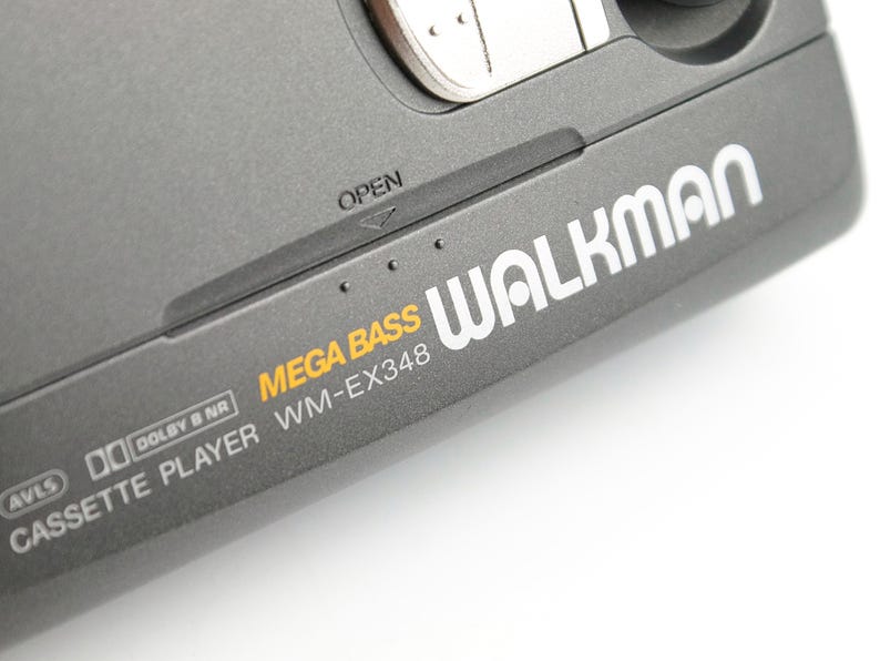 May include: A dark gray portable cassette player with the text WALKMAN, MEGA BASS, and WM-EX348 printed on the surface. The device features an OPEN latch, AVLS, and Dolby B NR branding, with a metallic silver button visible at the top edge.