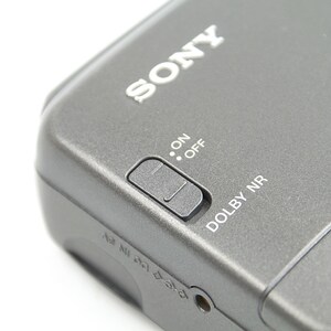May include: A dark gray Sony portable cassette player featuring a sliding switch labeled ON and OFF, with the text DOLBY NR printed nearby. The device includes a small circular port and additional small icons embossed on the side casing.