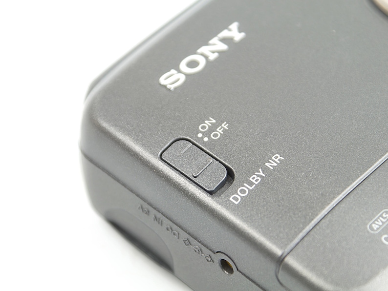 May include: A dark gray Sony portable cassette player featuring a sliding switch labeled ON and OFF, with the text DOLBY NR printed nearby. The device includes a small circular port and additional small icons embossed on the side casing.