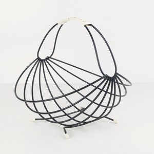 Op de afbeelding: Een zwarte metalen tijdschriftenrek met een witte handgreep en witte voetjes. Het rek is waaiervormig en heeft een gebogen ontwerp.