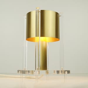 May include: A gold table lamp with a clear acrylic base and shade. The lamp is turned on and the light is shining through the shade.