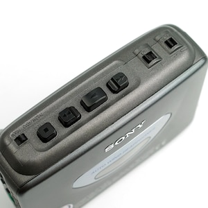 May include: A dark gray Sony portable cassette player featuring a top-mounted control panel with black playback buttons, a tape type selector switch labeled NORM, CRO2, METAL, and two input ports. The device has a silver oval accent on the top.
