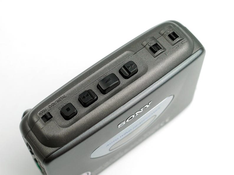May include: A dark gray Sony portable cassette player featuring a top-mounted control panel with black playback buttons, a tape type selector switch labeled NORM, CRO2, METAL, and two input ports. The device has a silver oval accent on the top.
