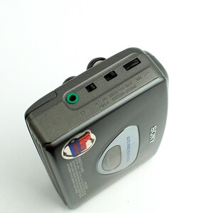 May include: A dark gray Sony portable cassette player featuring a green headphone jack, volume control wheel, and a small circular window. The device has a silver Sony logo and a small circular sticker with red and blue graphics on the top surface.