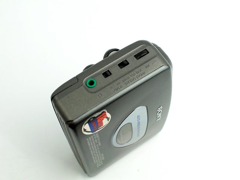 May include: A dark gray Sony portable cassette player featuring a green headphone jack, volume control wheel, and a small circular window. The device has a silver Sony logo and a small circular sticker with red and blue graphics on the top surface.