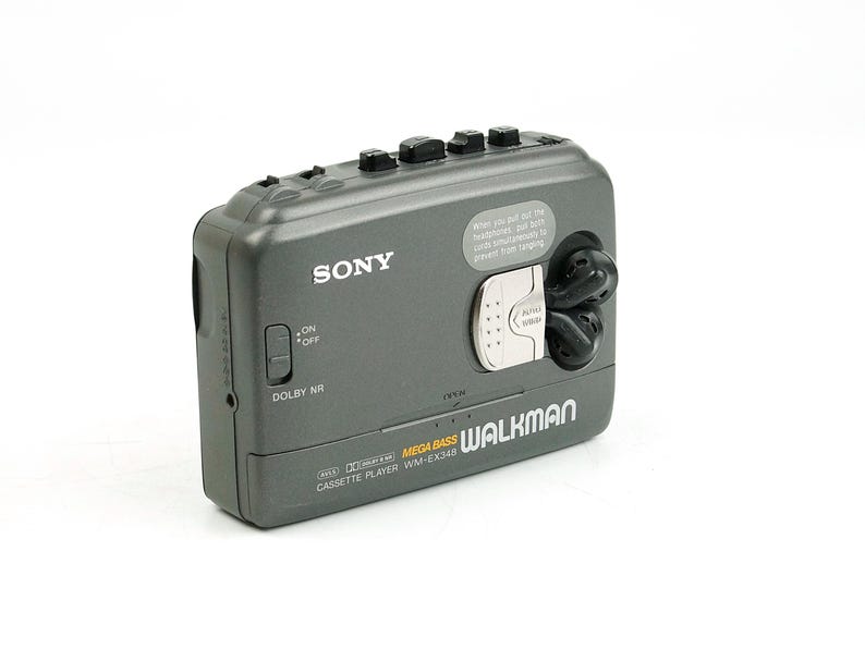 May include: A dark gray Sony Walkman cassette player model WM-EX348 featuring Mega Bass, Dolby NR, and an on/off switch. The device includes a silver latch, black control buttons on the top edge, and a small circular area for storing earbuds.